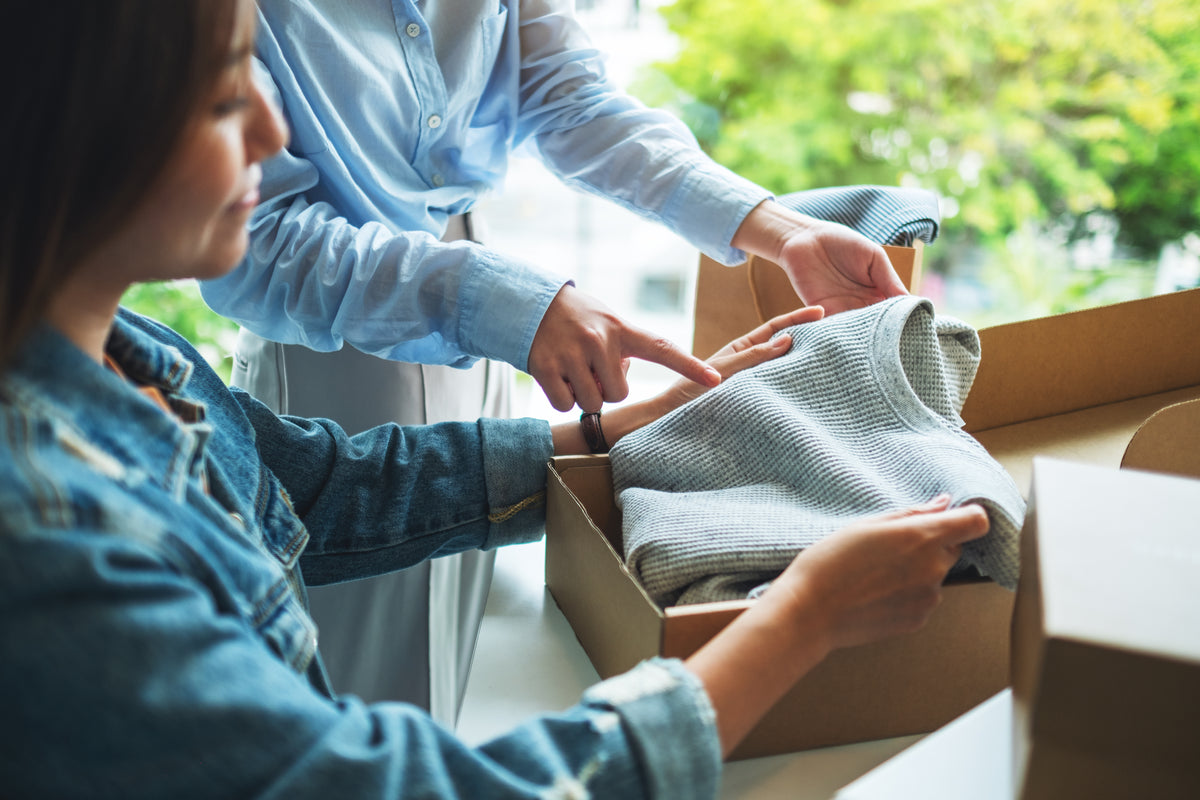 Two people unpacking a box of clothes in a bright room with greenery outside.